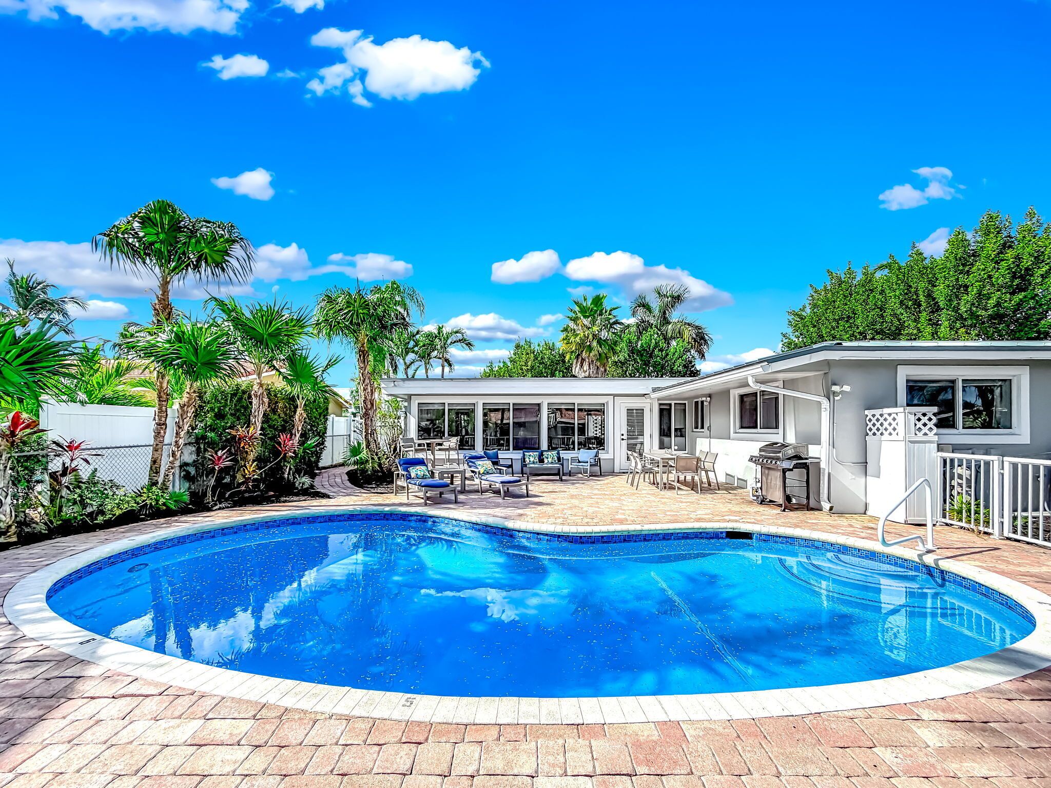 A swimming pool with a brick deck, surrounded by palm trees and a covered patio area with outdoor furniture.
