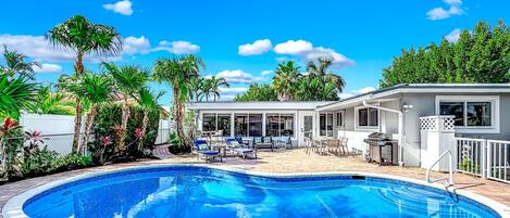 A swimming pool with a brick deck, surrounded by palm trees and a covered patio area with outdoor furniture.