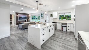 A modern kitchen with white cabinetry, a central island, and a large window providing a view of greenery. - Heated Pool! Kayak & Waterfront +1 Mile To Beach! (Pompano Beach)