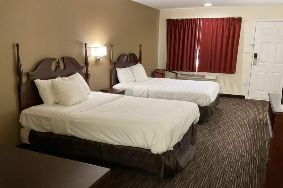 A hotel room with two beds, a desk, a chair, and a window with red curtains.