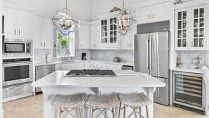 A modern kitchen with white cabinetry, stainless steel appliances, a marble island, and wicker bar stools.