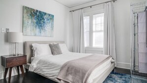 Een slaapkamer met een bed, nachtkastje, schilderij, raam met gordijnen en een douchegedeelte.