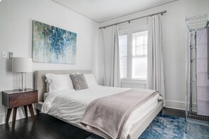 A bedroom with a bed, a nightstand, a painting, a window with curtains, and a shower area. - Historic Candler Park Gem! -Sleeps 4- Apt 3 - AC Atlanta (Atlanta)