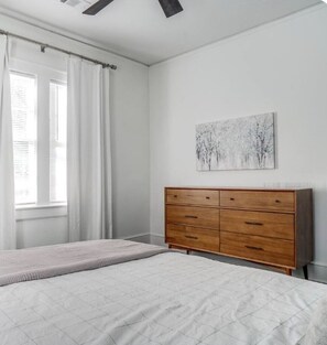 Een slaapkamer met een bed, een houten dressoir, een raam met gordijnen en een schilderij aan de muur.