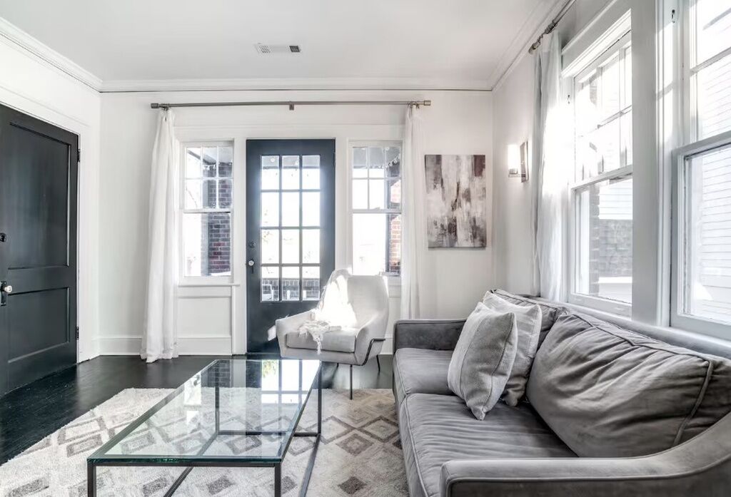 A living room with a glass coffee table, a grey sofa, a white armchair, and a dark door.