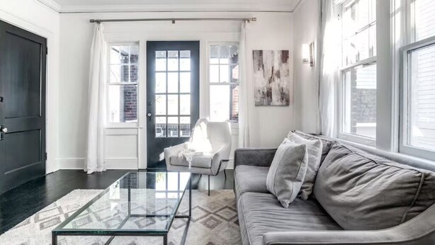 Een woonkamer met een glazen salontafel, een grijze bank, een witte fauteuil en een donkere deur.