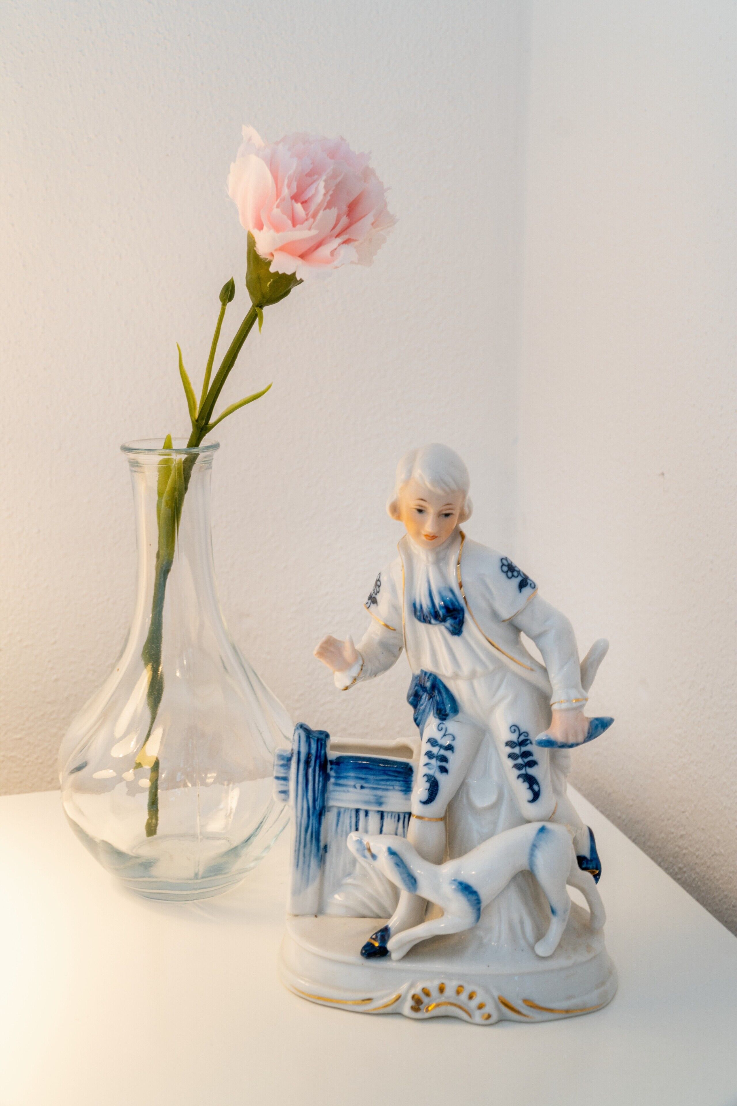 Eine transparente Glasvase mit einer rosa Blume und einer Keramikfigur einer Person mit einem Hund.
