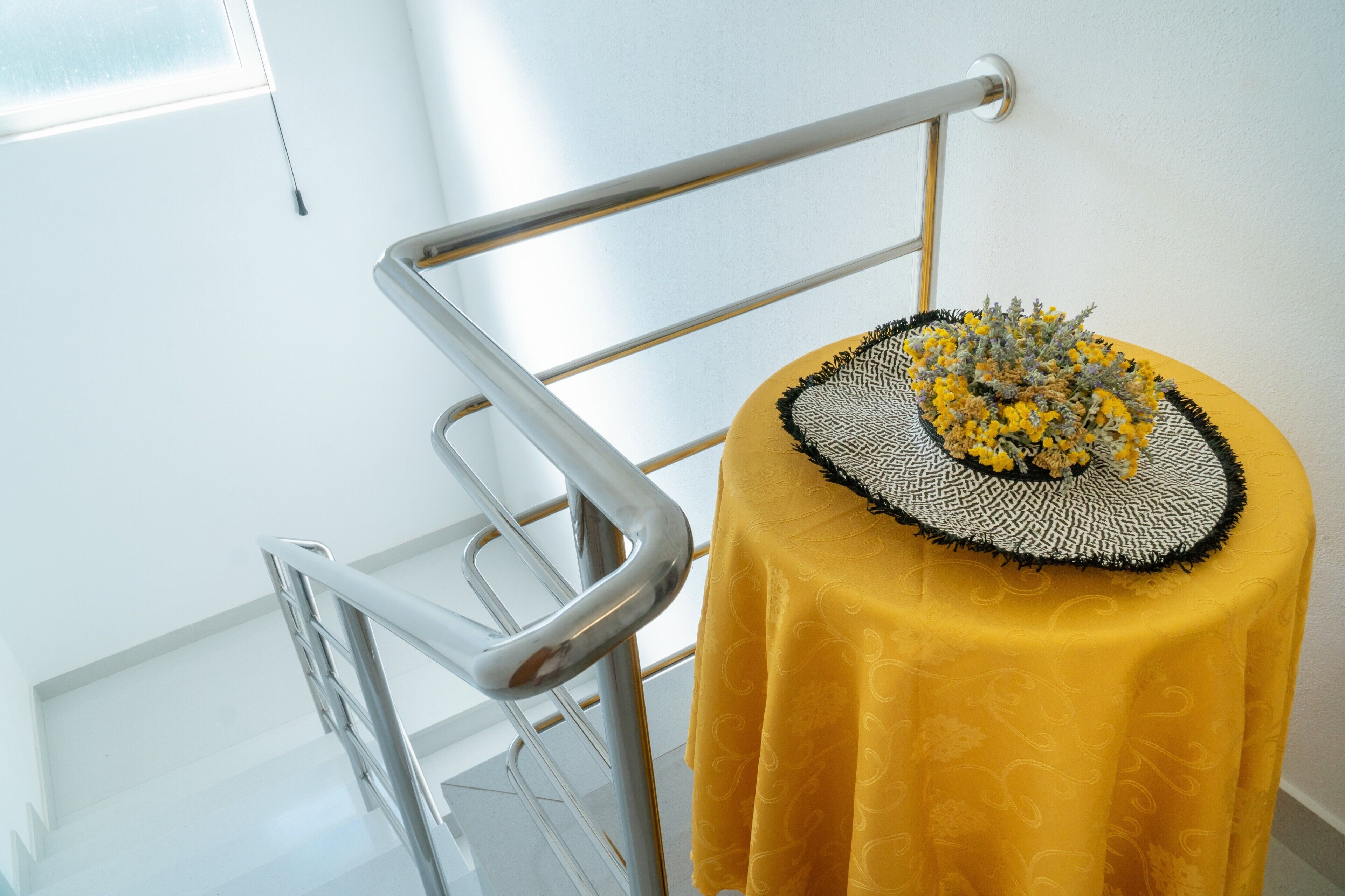 A small round table with a yellow tablecloth and a decorative centerpiece, positioned next to a metal railing in a bright room with white walls.