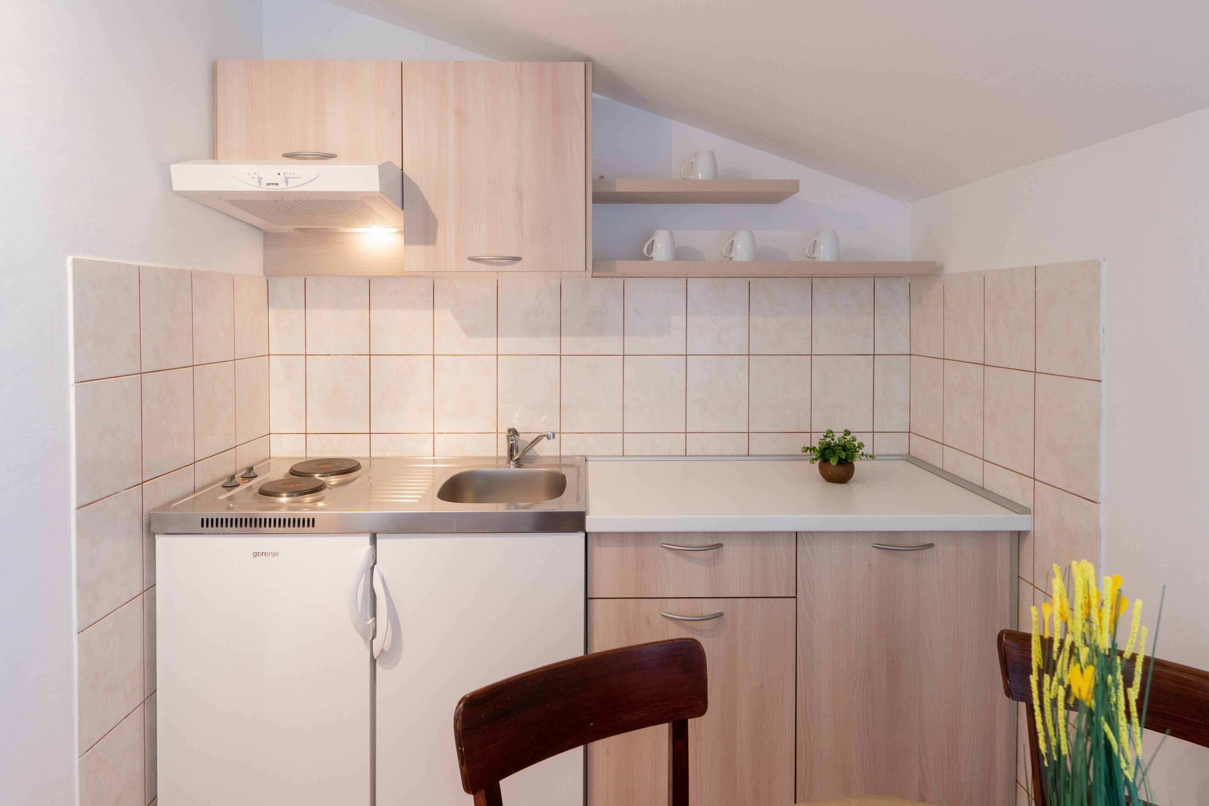 A compact kitchen with a built-in oven, a sink, and a small countertop. There is a wooden chair and a potted plant on the counter.