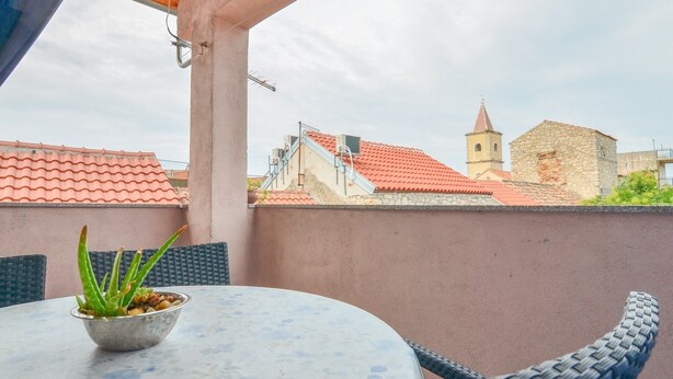 A balcony with a table and chairs, a potted plant, and a view of buildings with red-tiled roofs and a church tower. - Apartment with old town view no.2 (Pirovac)