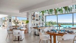 Un comedor amplio con sillas blancas y mesas de madera, rodeado de ventanas grandes que ofrecen vistas al océano.