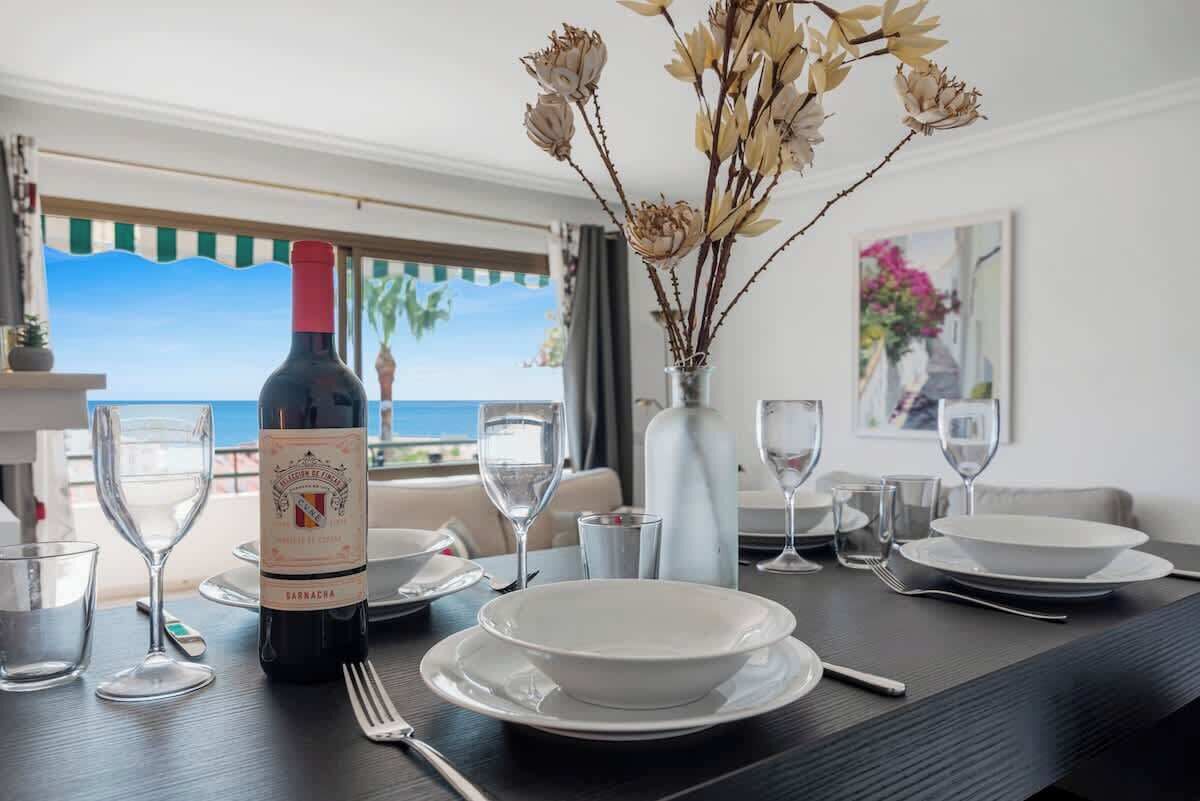 A dining table set with white plates, wine glasses, and a bottle of wine. There is a vase with dried flowers on the table.