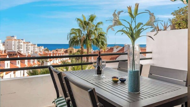 Ein Balkon mit Tisch, Stühlen, einer Flasche und Gläsern. Man sieht auf das Meer und Palmen.