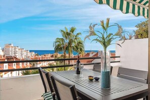 Un balcon avec une table, des chaises, une bouteille et des verres. On peut admirer la mer et les palmiers.