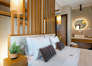 A modern bedroom with a wooden headboard, a bedside table with a plant, a large bed with white linens, and a bathroom with a round mirror and wooden vanity. - Terra Lucis Mountain Residences Anima Villa (Zakynthos)