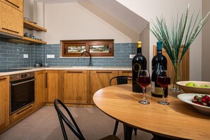 A kitchen with wooden cabinets, a built-in oven, a window, and a dining table with wine glasses and a bowl of fruit. - Terra Lucis Mountain Residences Anima Villa (Zakynthos)