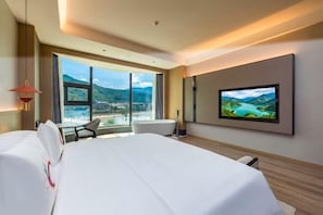 Une chambre d’hôtel avec un grand lit, un téléviseur fixé au mur, une chaise et une vue sur un paysage montagneux par de grandes fenêtres.