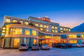 A multi-story hotel with a curved facade, large windows, and a parking lot with several cars. - Jiuzhou Jindu Hotel (Pingwu)