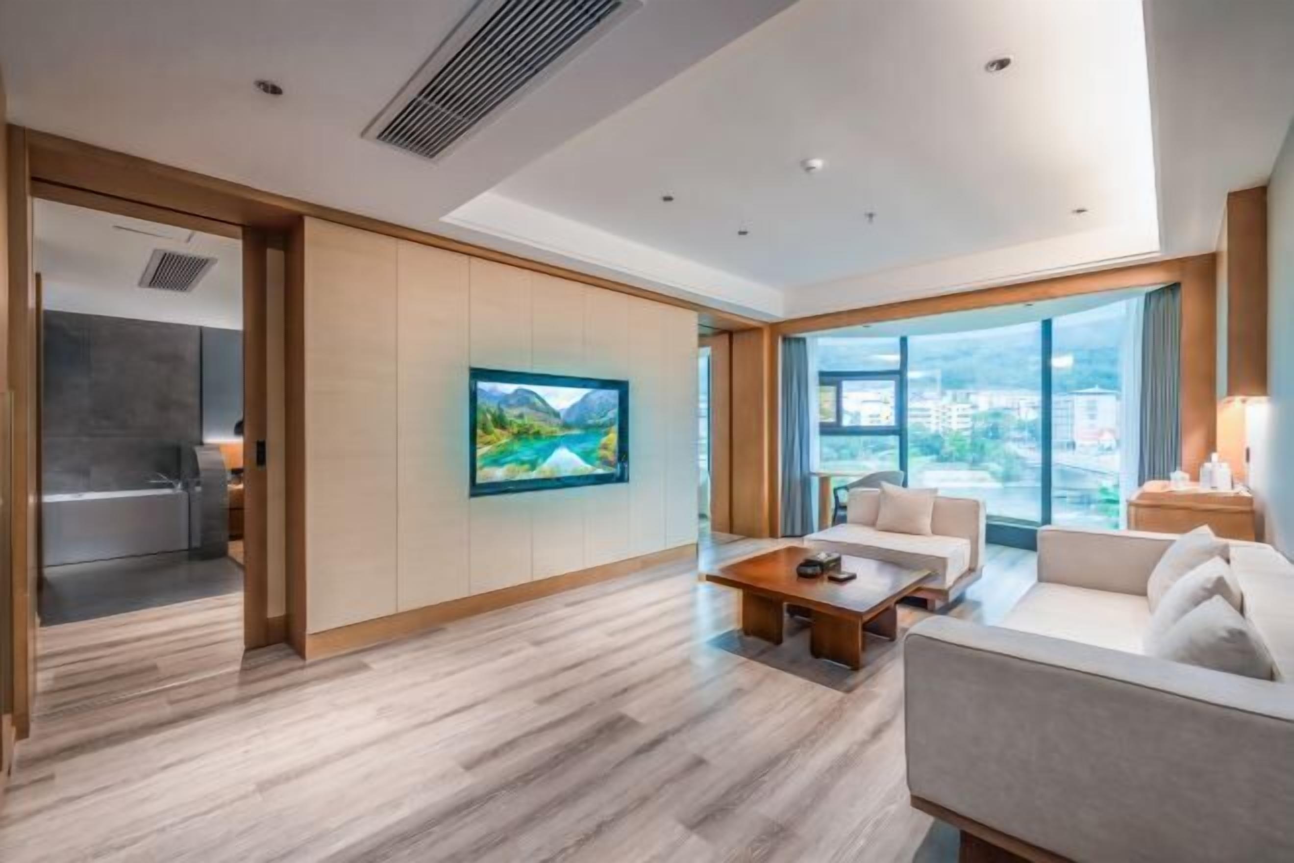 Une chambre d’hôtel moderne avec un plancher en bois, une grande fenêtre, un canapé, une table basse et un tableau au mur.