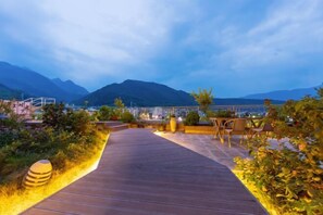 Un sentier en bois mène à une terrasse offrant une vue sur la montagne, avec des sièges d’extérieur et une rambarde.
