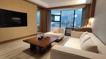 A modern hotel room with a large window offering a city view, a flat-screen TV mounted on the wall, and a comfortable seating area with a sofa and a coffee table.