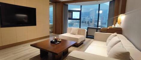 A modern hotel room with a large window offering a city view, a flat-screen TV mounted on the wall, and a comfortable seating area with a sofa and a coffee table.