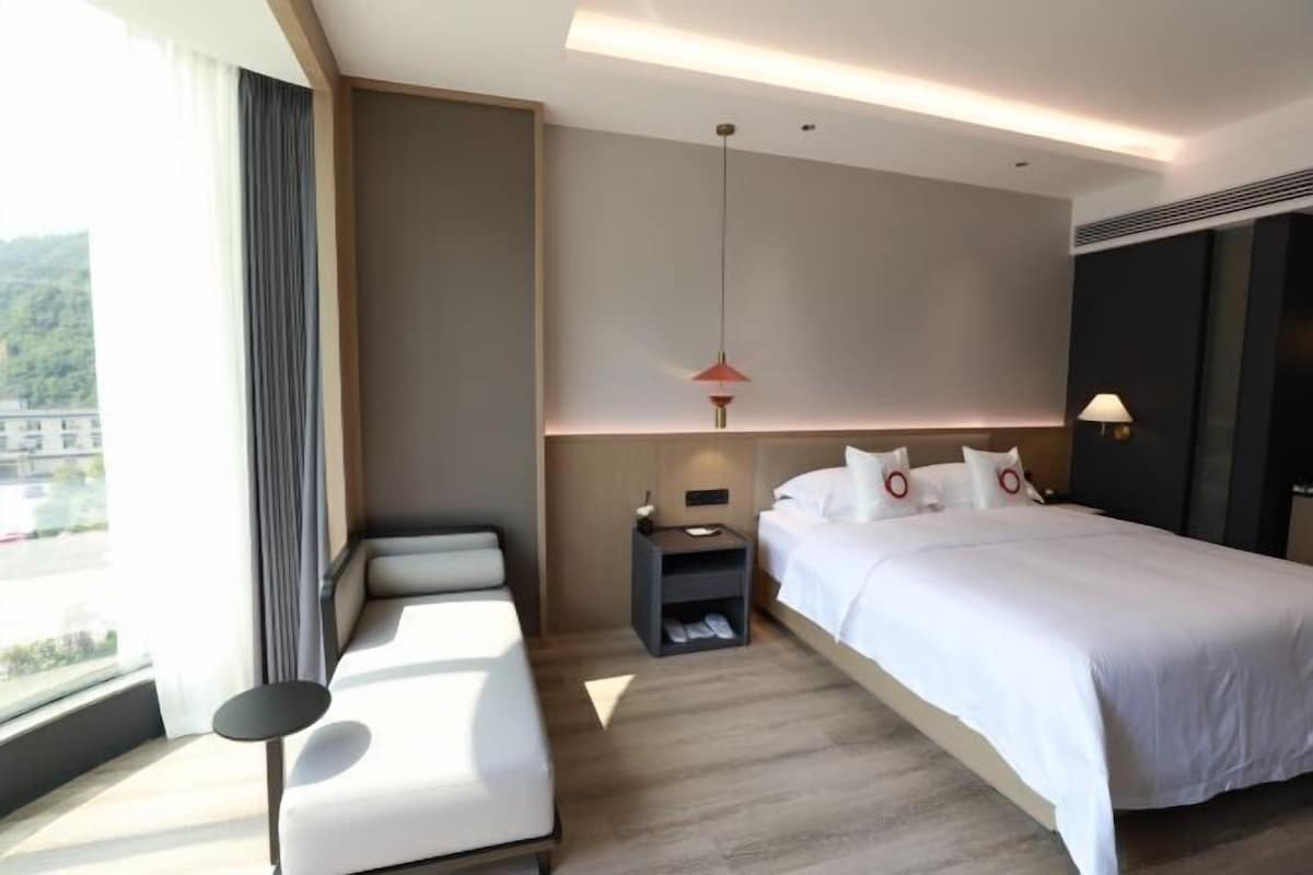 Een moderne hotelkamer met een groot bed, een sofa, een nachtkastje en uitzicht op het groen.