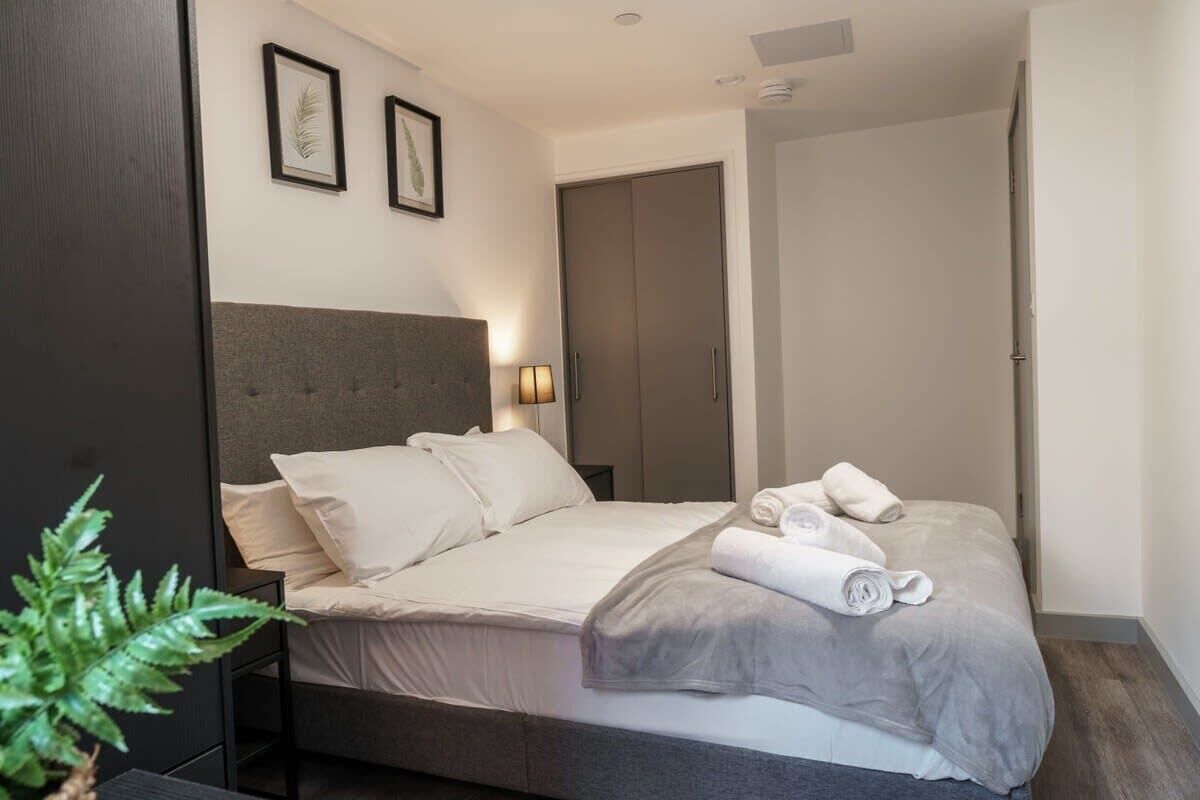 A modern hotel room with a large bed, a bedside table, a wall-mounted lamp, and a door leading to another room.