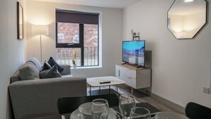 A modern living room with a sofa, a dining table with chairs, a TV, and a large window with a view of a brick building. - Contemporary 1BR Apt in Liverpool near City Centre (Liverpool)