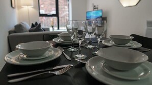 A dining table set with white plates, bowls, cutlery, and wine glasses, with a TV and window in the background. - Contemporary 1BR Apt in Liverpool near City Centre (Liverpool)