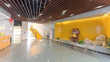 A modern lobby with a wooden ceiling, a reception desk, a yellow wall with a bear decoration, and a staircase in the background.