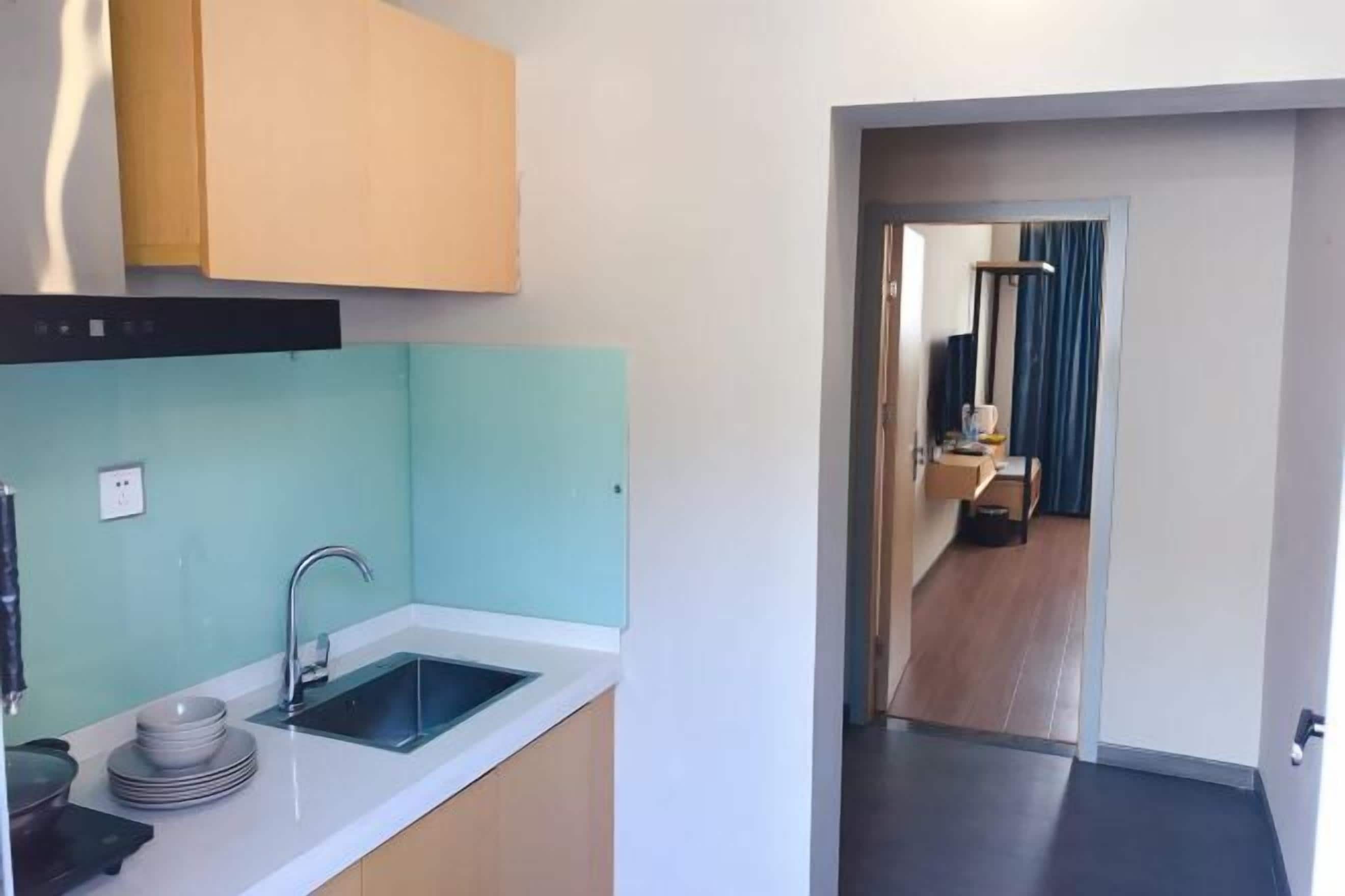 A modern kitchen with a sink, cabinets, and a countertop. There is a door leading to a room with a mirror and a bed.