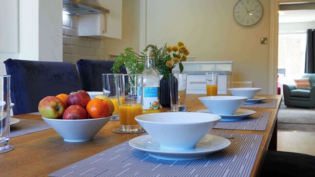 A breakfast setting with a fruit bowl, glasses, and a bottle on a wooden table.