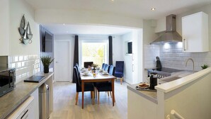A modern kitchen with white cabinetry, a stainless steel range hood, and a built-in oven. There is a dining area with a wooden table and blue chairs.