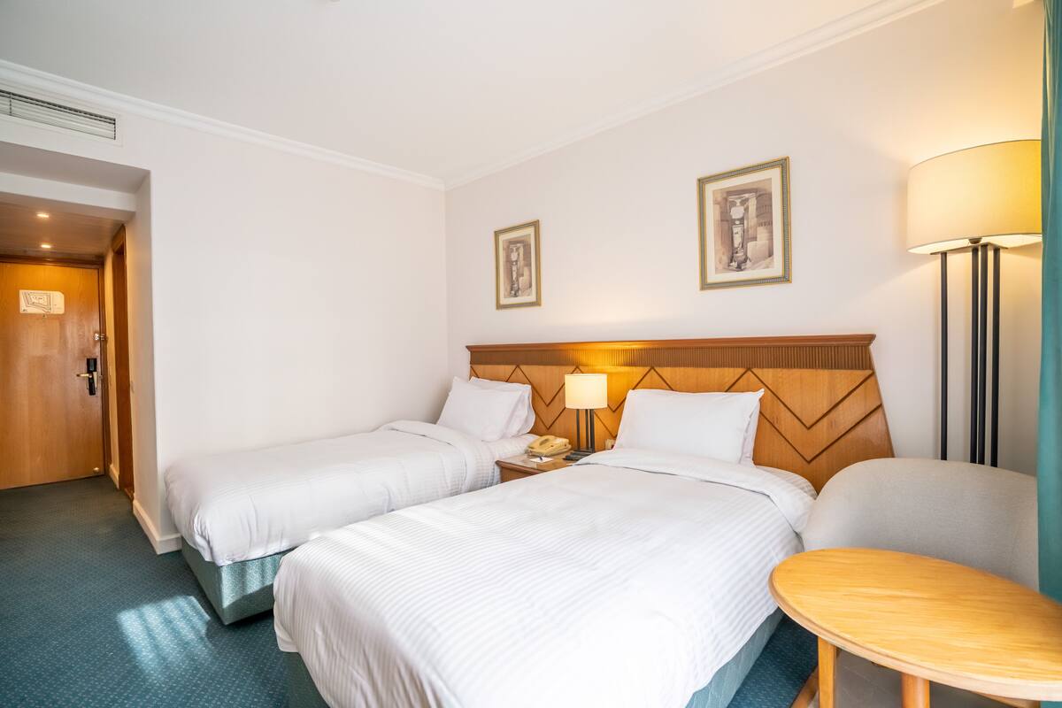 A hotel room with two beds, a wooden headboard, a bedside table with a lamp, and a small round table.