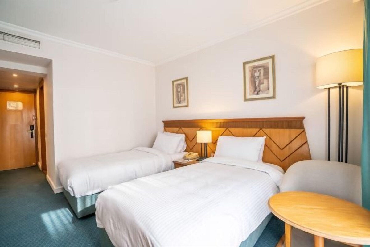Habitació d'hotel amb dos llits, un panell de capçalera de fusta, una taula de nit amb una làmpada i una taula rodona petita.