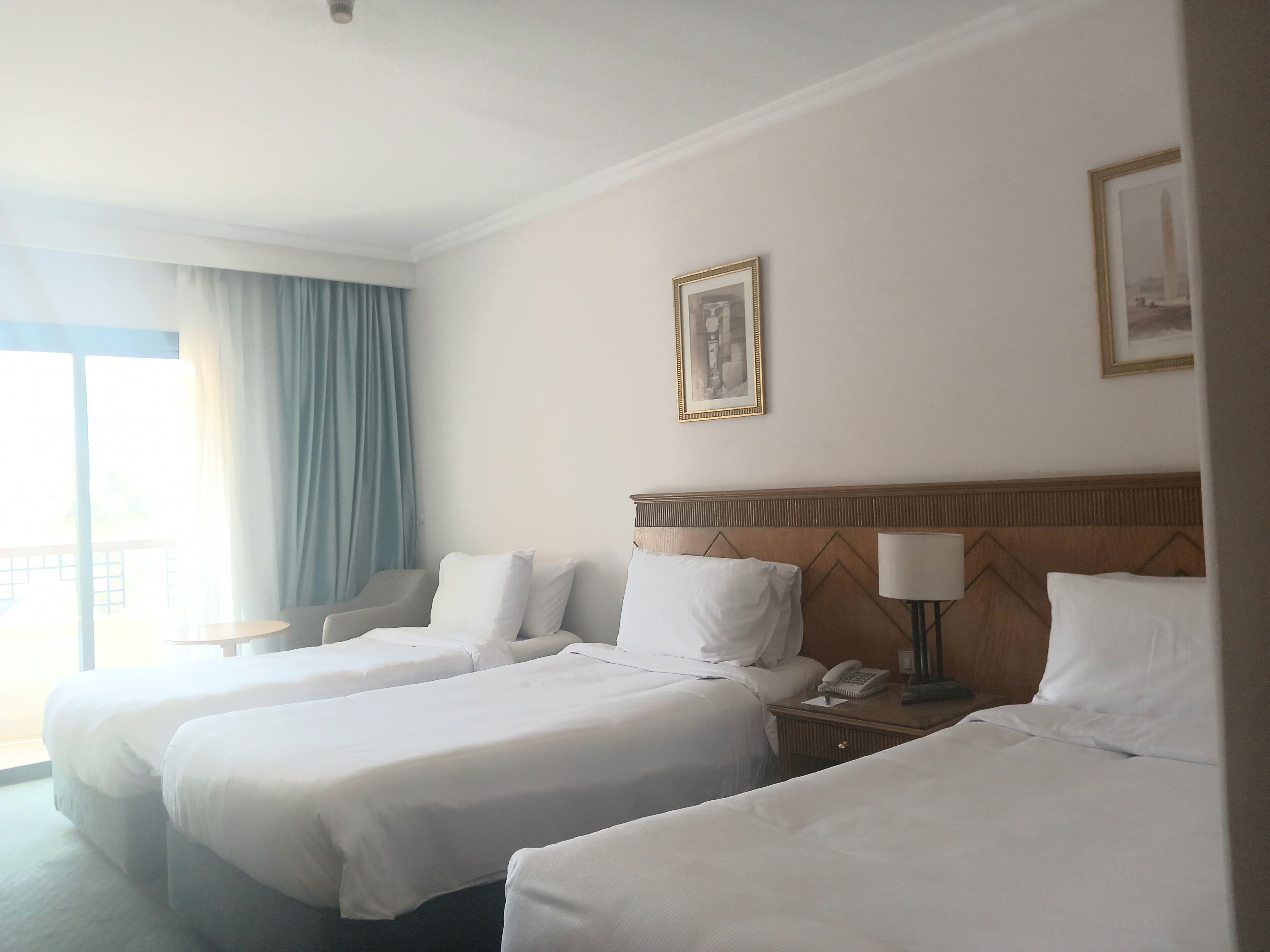 Una habitación de hotel con dos camas, cabecera de madera, mesita de noche con lámpara y un cuadro en la pared.