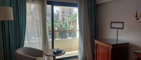 Quarto de hotel com cama, uma cadeira, uma mesinha com caixa de lenços, escrivaninha e vista para edifícios e palmeiras pela janela.