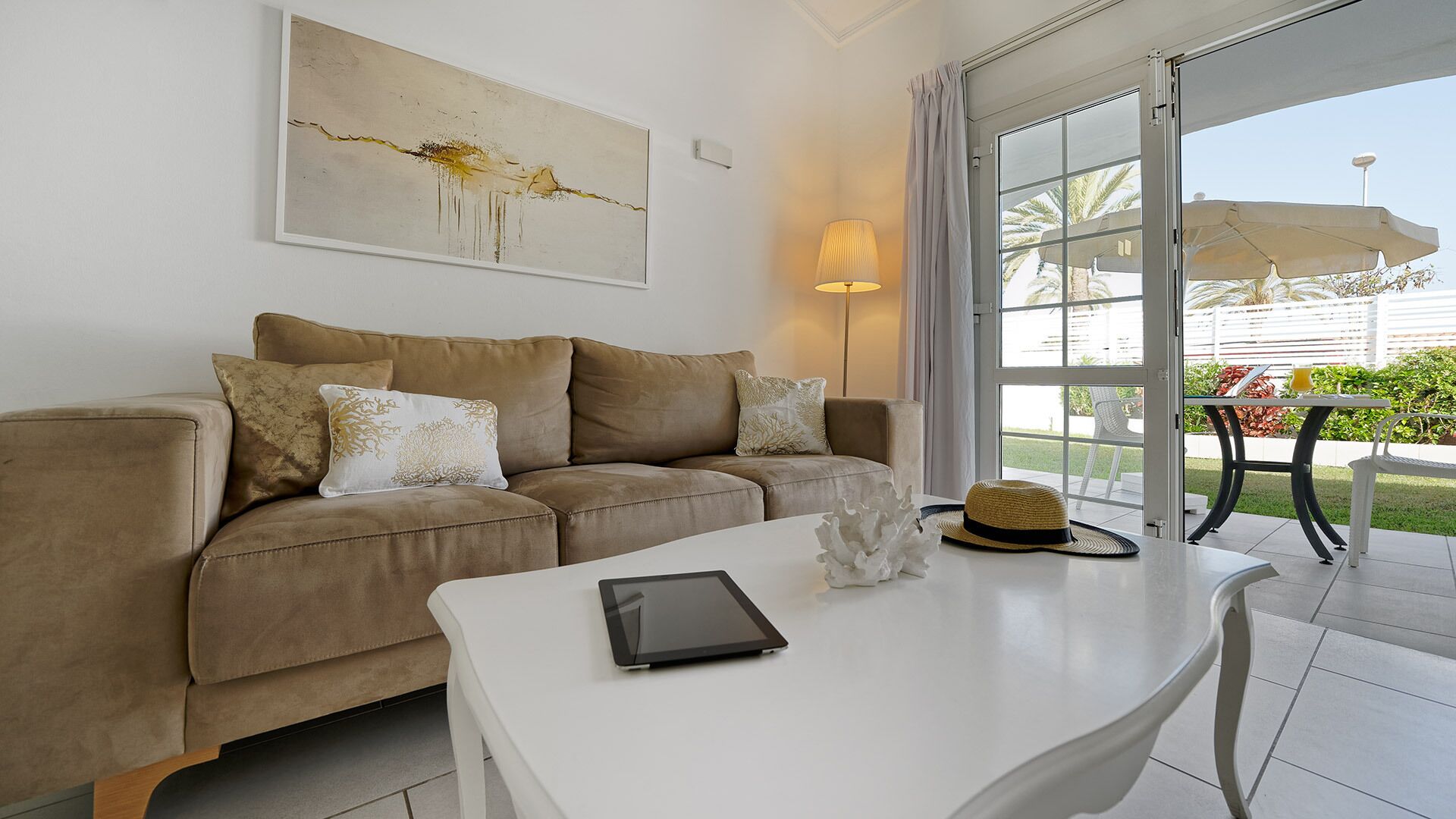Ein modernes Wohnzimmer mit einem beige gefärbten Sofa, einem weißen Couchtisch und einem großen Fenster mit Blick ins Freie.