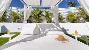 A white canopy bed with a tablet and a straw hat on the bed, two white nightstands with drinks, and a white fence with palm trees in the background. - Bungalows Adonis (San Bartolomé de Tirajana)
