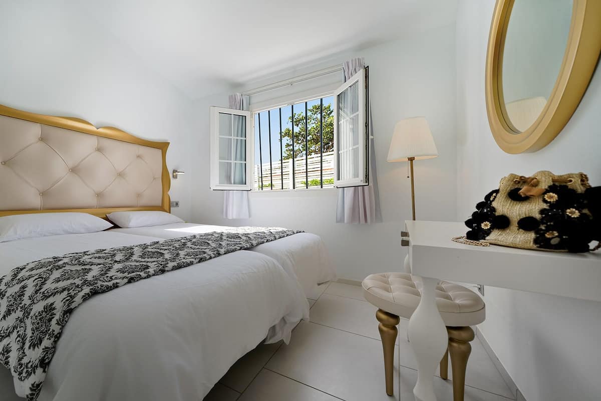 A bedroom with a large bed, a bedside table, a lamp, a mirror, and a view of the outdoors.