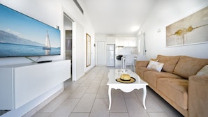 A modern living room with a flat-screen TV, a sofa, a coffee table, and a kitchen in the background. - Bungalows Adonis (San Bartolomé de Tirajana)