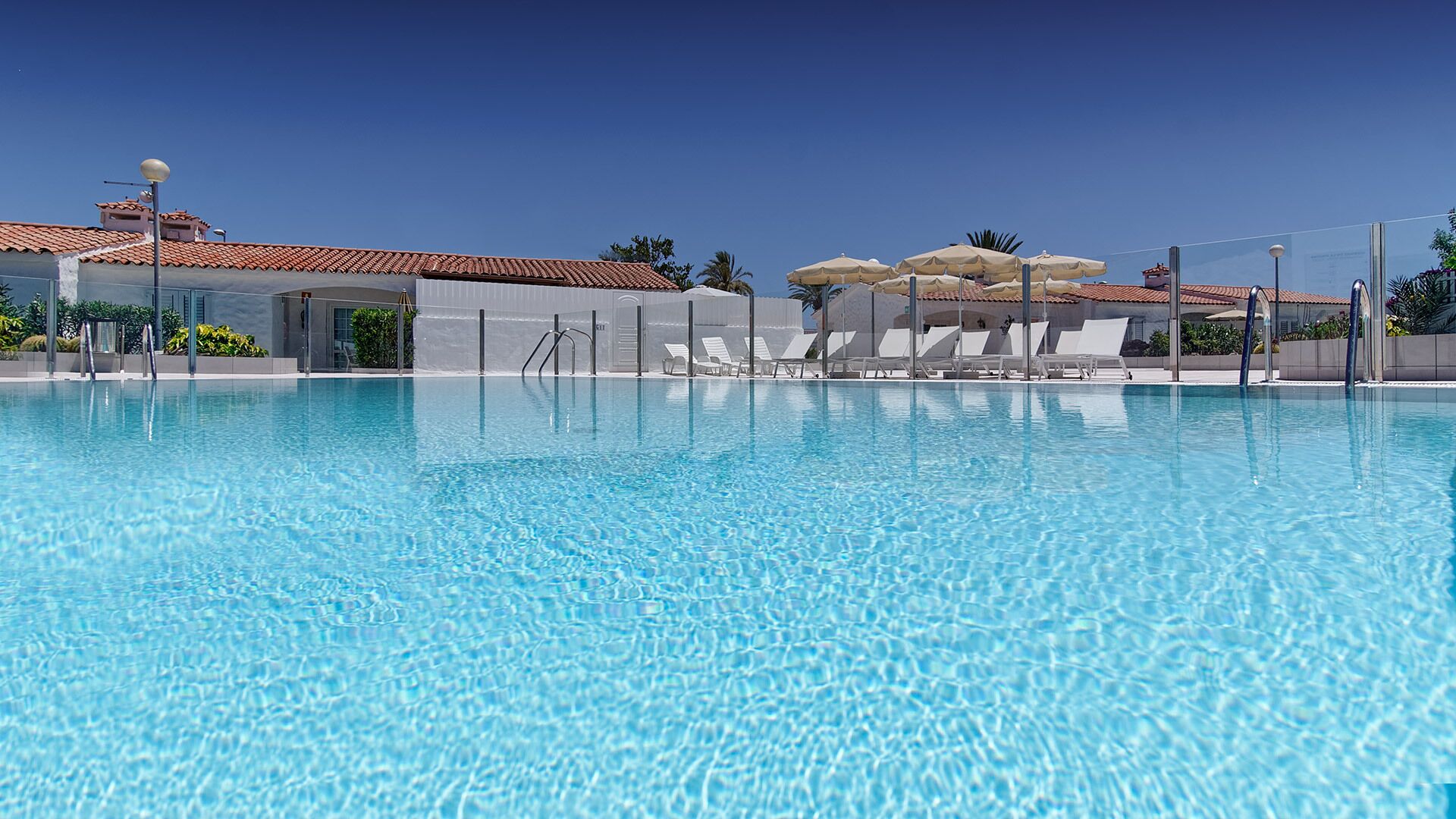 A clear blue pool with white lounge chairs and umbrellas, surrounded by buildings with red-tiled roofs.