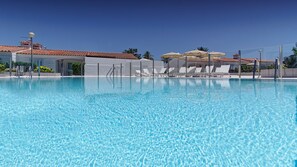 A clear blue pool with white lounge chairs and umbrellas, surrounded by buildings with red-tiled roofs. - Bungalows Adonis (San Bartolomé de Tirajana)