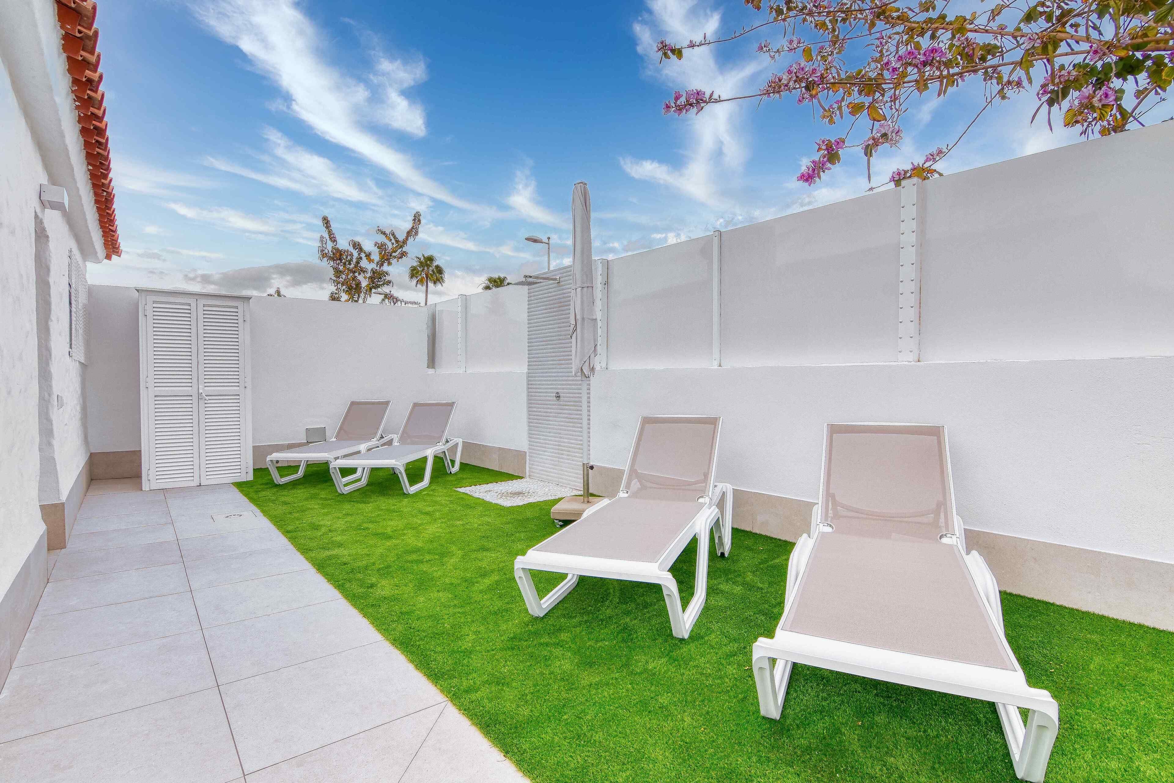 A patio with white lounge chairs and artificial grass, surrounded by white walls and a clear sky.