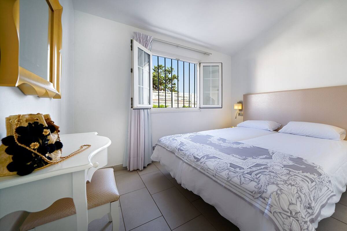 A hotel room with a large bed, a desk with a chair, a bedside table with a lamp, and a window with a view of buildings.
