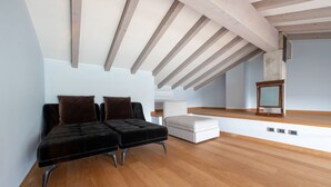 A spacious loft bedroom with exposed wooden beams, a black upholstered bench, a white ottoman, and a wooden dresser.