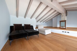 Une chambre spacieuse aménagée en mezzanine, avec des poutres apparentes, un banc recouvert de tissu noir, un pouf blanc et une commode en bois.