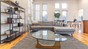 A modern living room with a sectional sofa, a glass coffee table, a black metal bookshelf, and large windows. - Stylish & Perfectly Located Deluxe 2-Bed Townhome (Dallas)