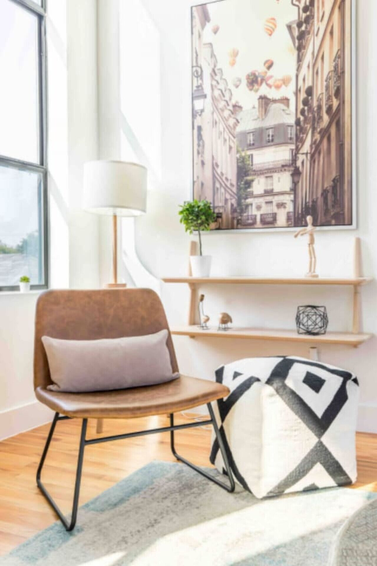 Una sala moderna con un sillón de cuero marrón, una lámpara de pie blanca, un cojín decorativo blanco y negro, y una pintura de un paisaje urbano en la pared.
